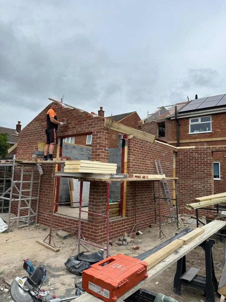 A construction worker stands on scaffolding while building a brick extension to a house, where sash window repair South London services may soon be needed. Wooden beams and building materials are scattered around the site.