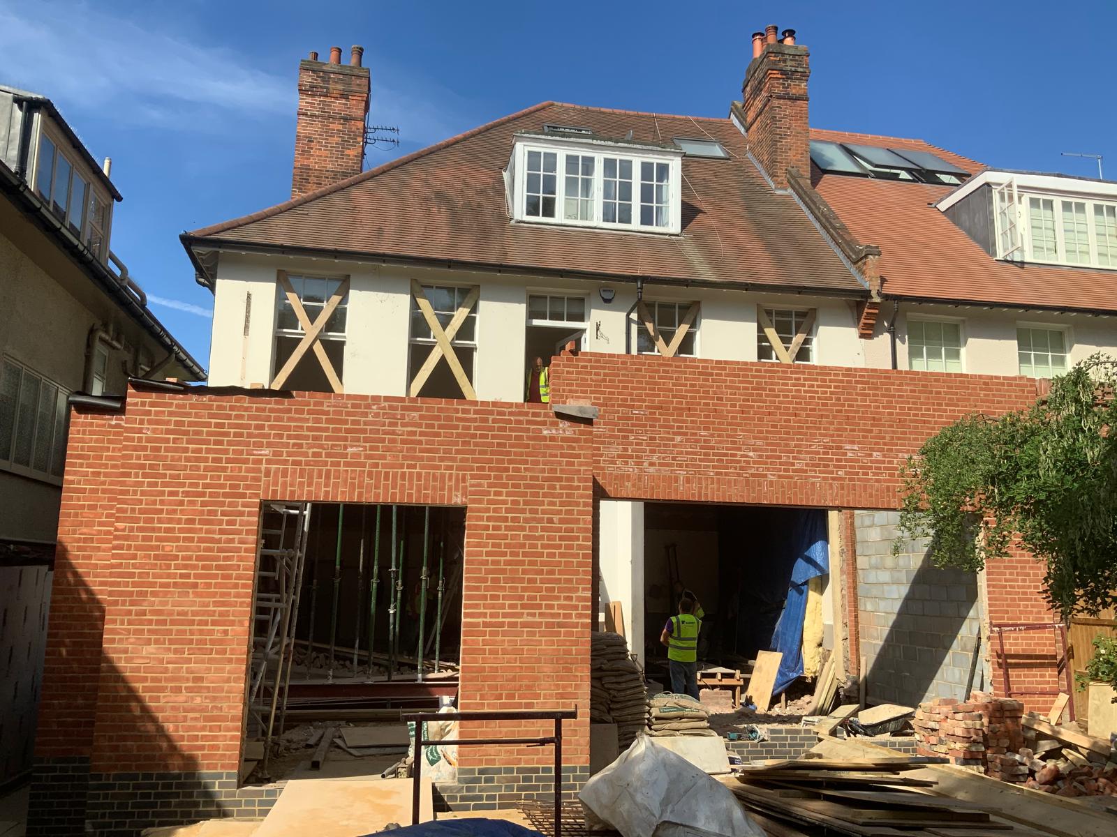 A brick extension under construction at the rear of a large house, with exposed beams and building materials visible; workers wearing high-visibility vests on site.