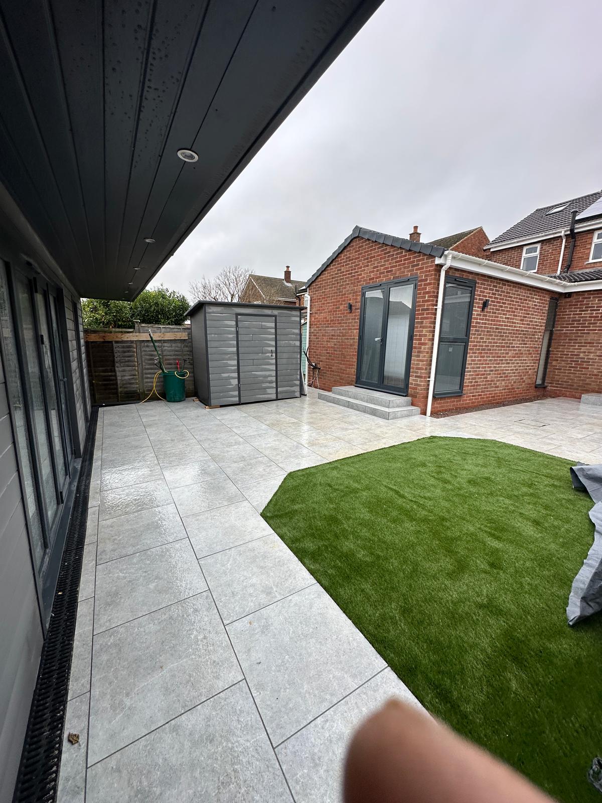 Modern back garden with large grey patio tiles, artificial grass area, a grey storage shed, and a red brick house on a cloudy day.