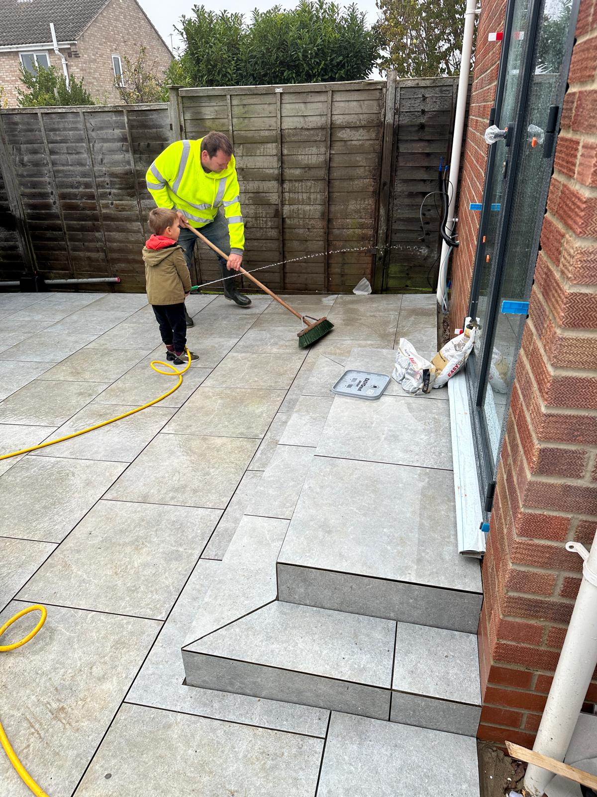 An adult and a child sweep newly laid patio tiles near the steps of a house; a yellow hose lies on the ground nearby.