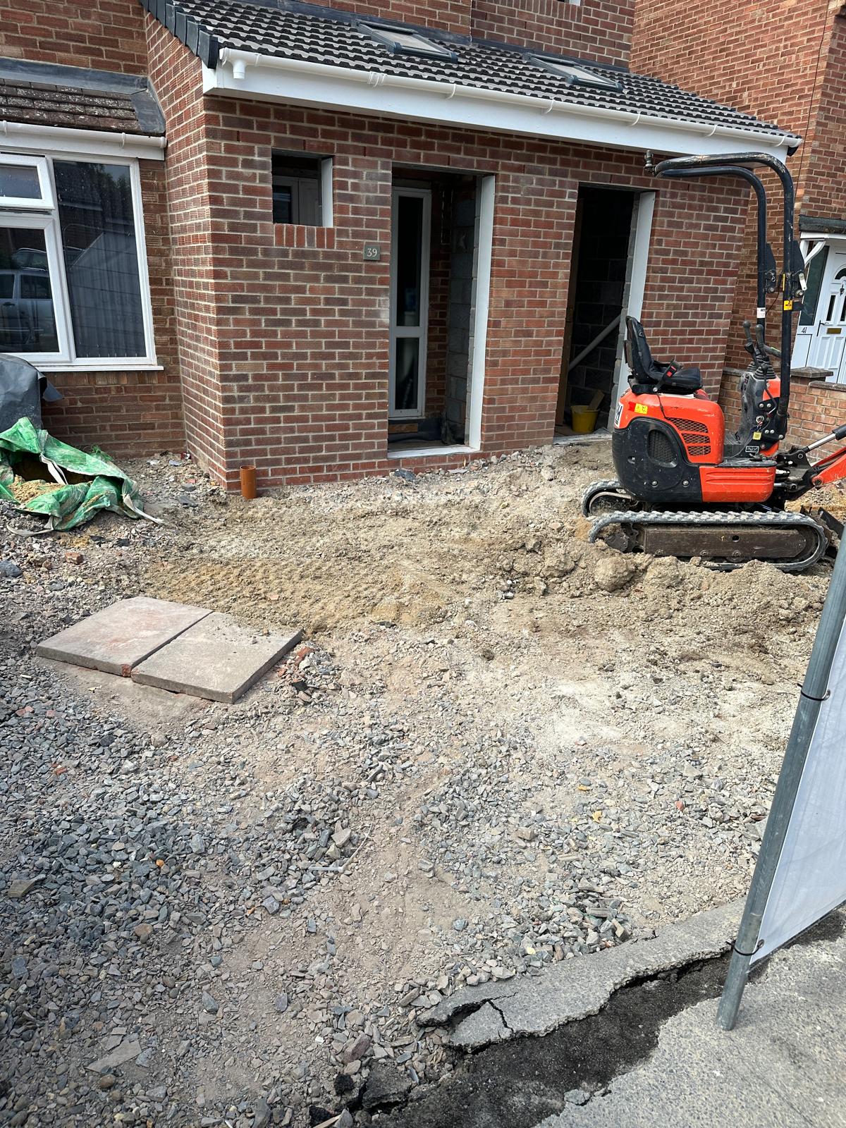 A small digger is parked on a gravel and dirt area in front of a brick house under construction or renovation, with building materials nearby.