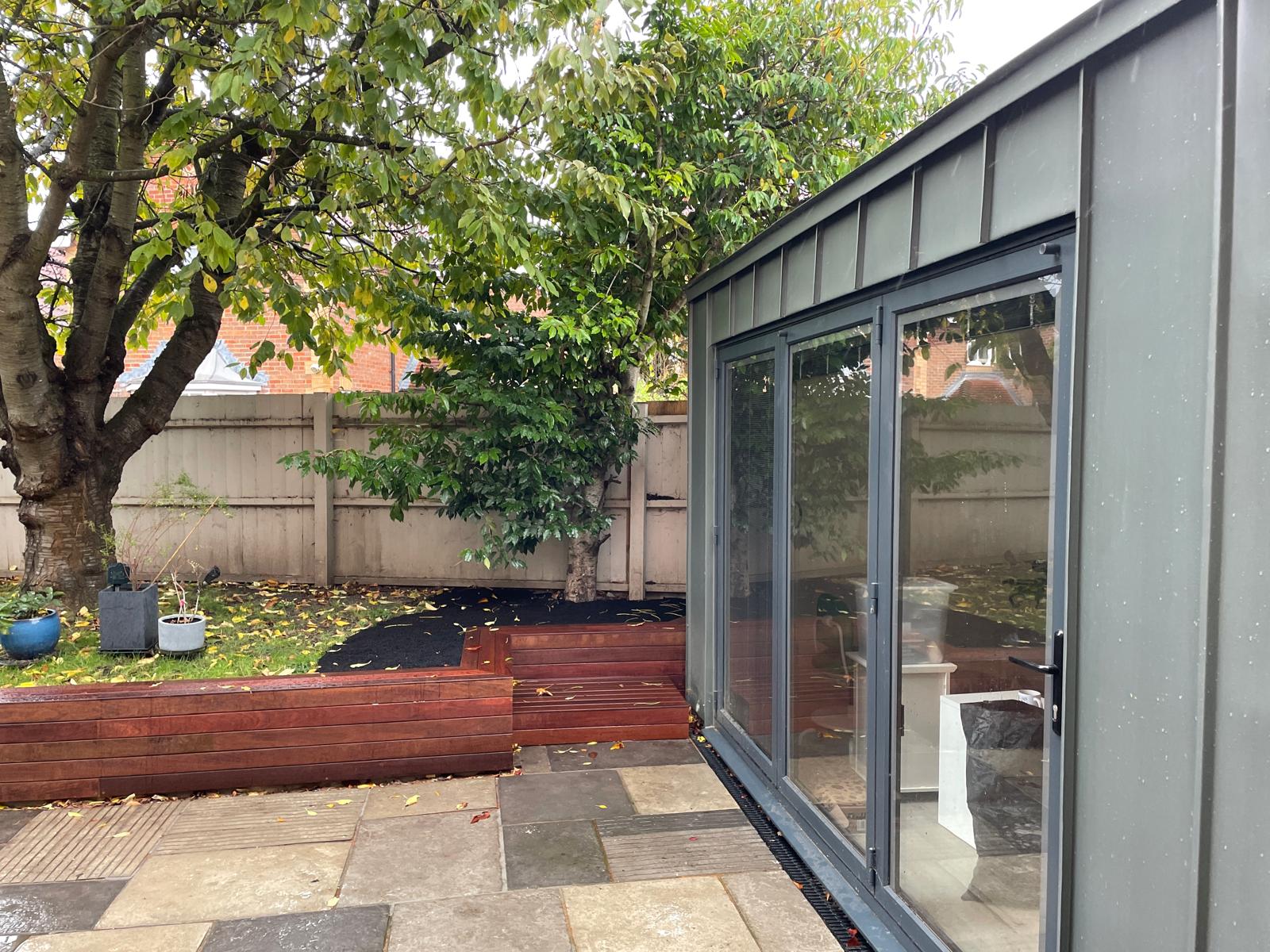 A modern garden room with large glass doors overlooks a paved patio and wooden planter beds, with trees and a fence in the background.