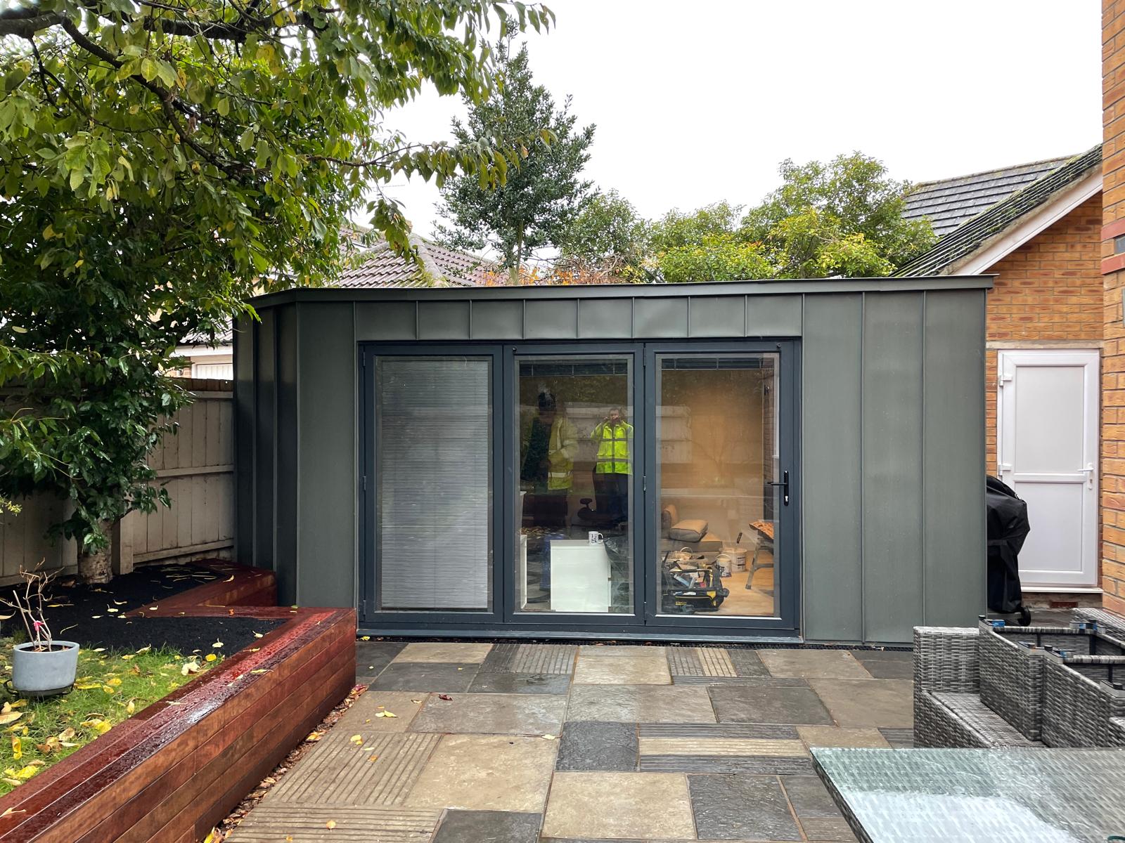 A modern garden office with large glass doors stands on a paved patio, surrounded by trees, plants, and outdoor furniture.