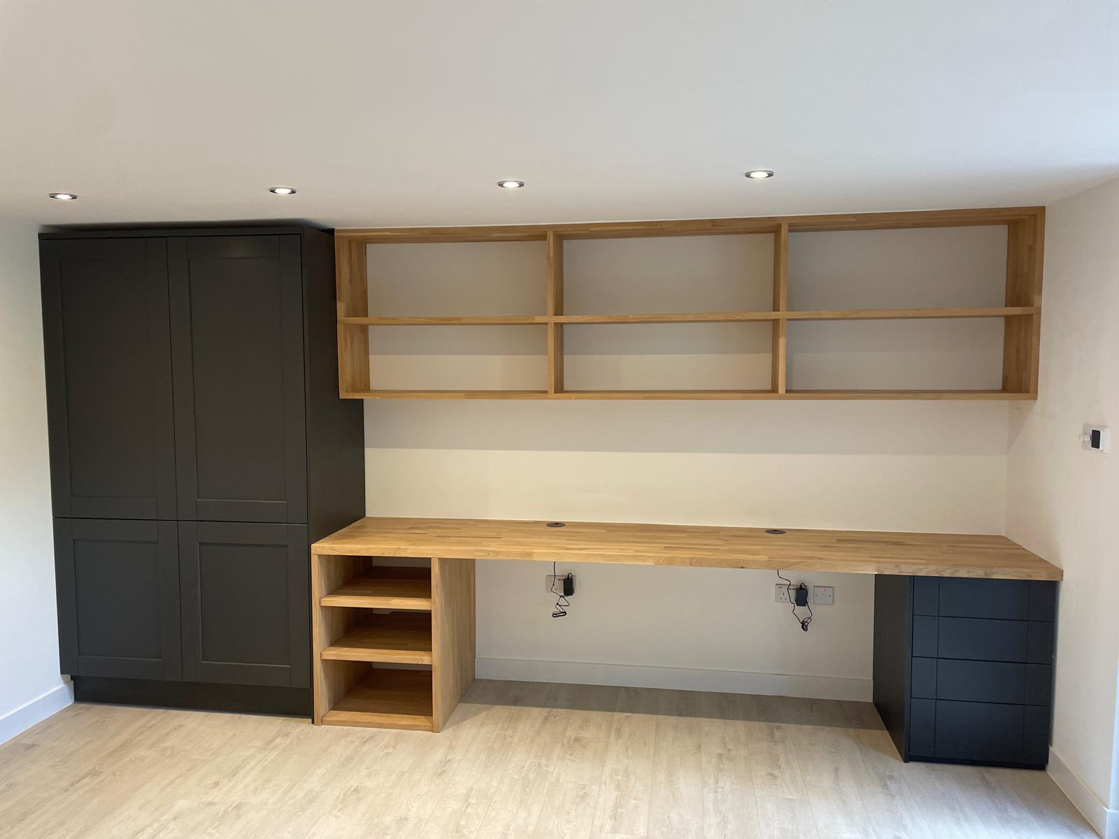 Minimalist home office setup with a wooden desk, open shelving, built-in black cupboard on the left, and black drawers on the right, against a white wall and light wood floor.