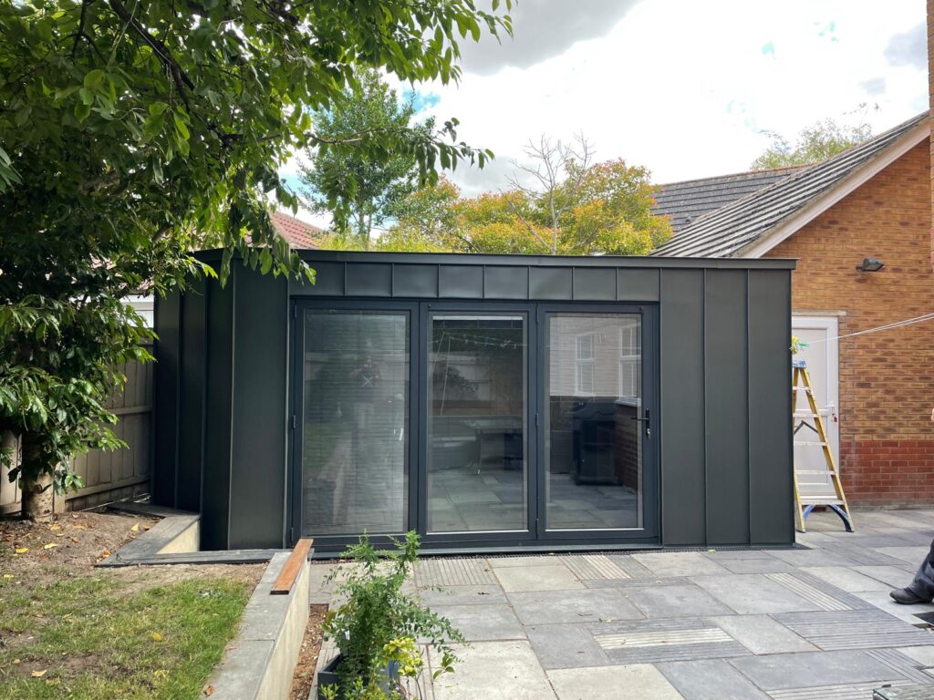 A modern black garden room with large sliding glass doors, situated on a patio next to a brick house and surrounded by trees.