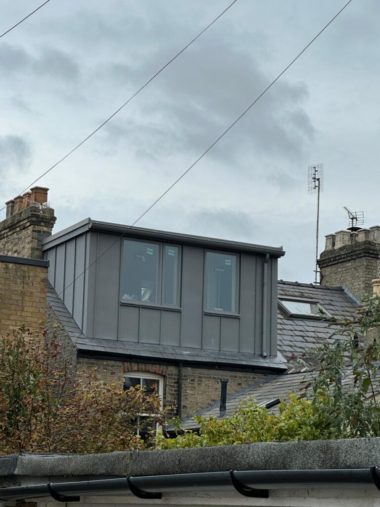 A modern grey dormer extension with large windows added to the roof of a brick house, surrounded by chimneys, plants, and overhead wires.