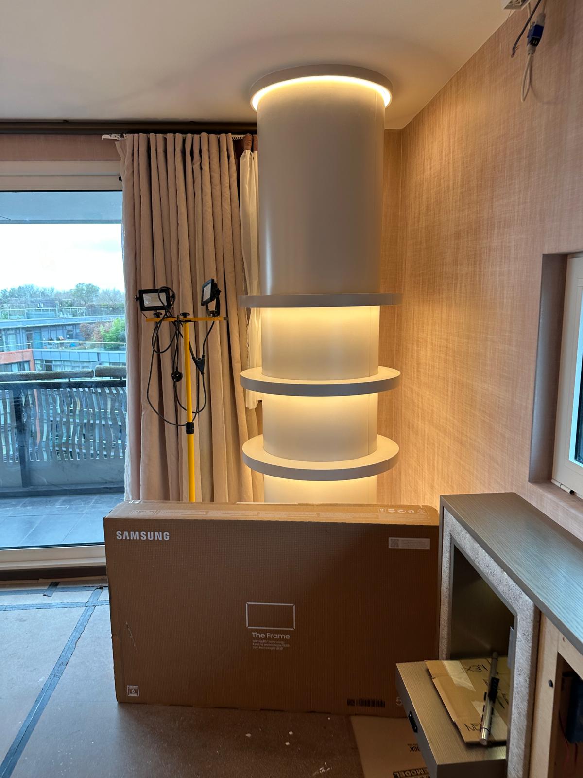 A modern interior with a cylindrical, backlit shelving unit, a covered window with curtains, a Samsung TV box, and a yellow work light in a partially furnished room.