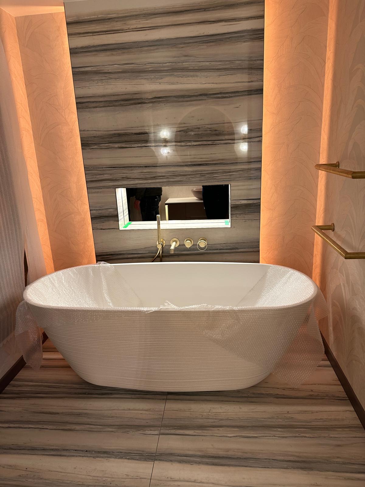 A modern bathroom with a freestanding white bath partially covered in bubble wrap, set against a striped marble wall with built-in shelf and gold fittings.