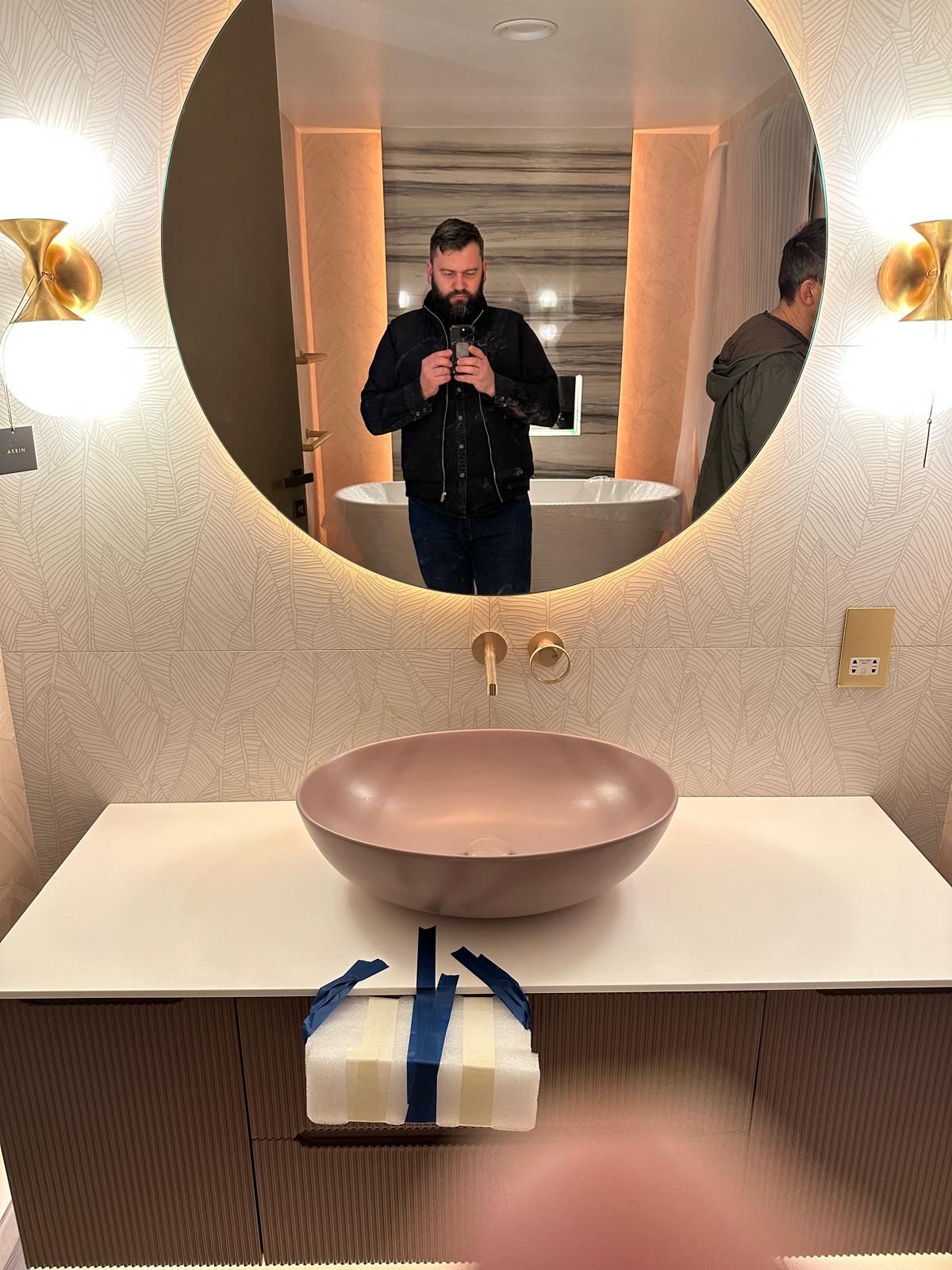 A man takes a photo of himself in a round mirror above a modern bathroom basin; another person is partially visible to the right.