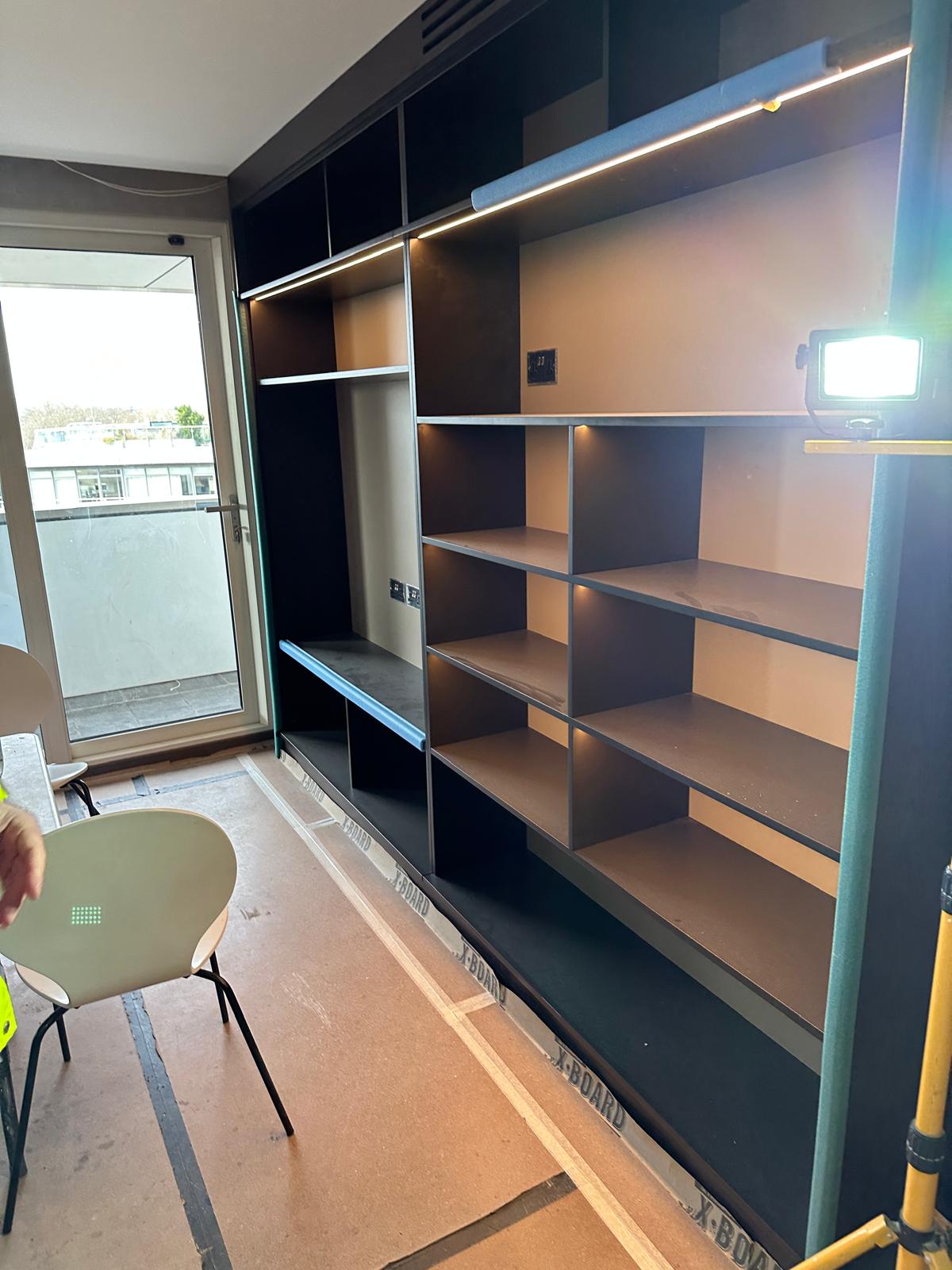 Open shelving and storage units installed along a wall in a modern room under construction, with a work light illuminating the space and a window in the background.