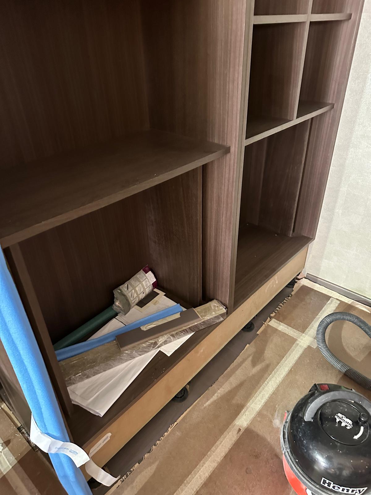 A wooden shelving unit with documents, tools, and materials on a lower shelf; a vacuum cleaner is on the floor nearby.