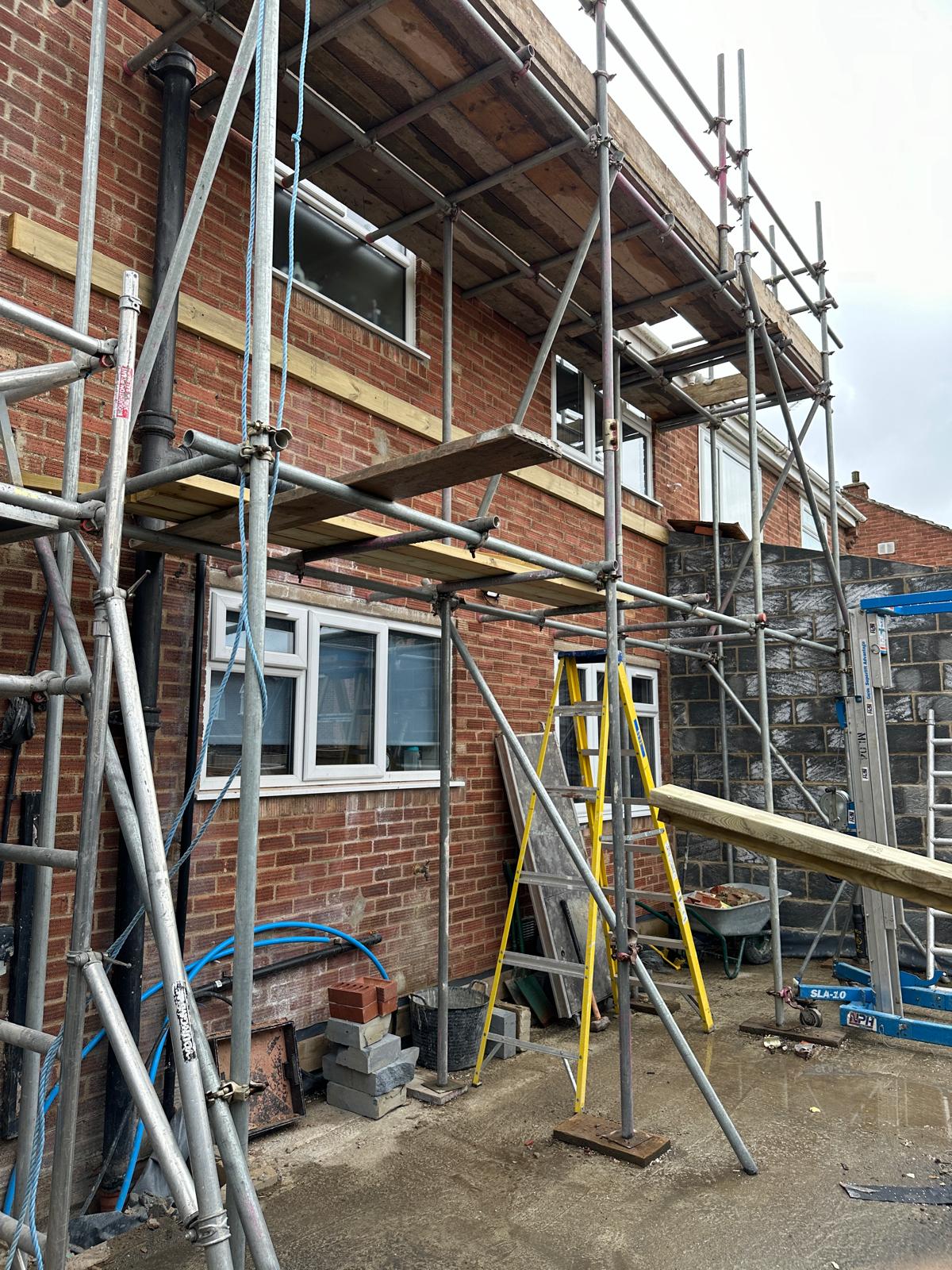 A brick house under construction with scaffolding, ladders, building materials, and tools visible; windows are fitted on both floors.