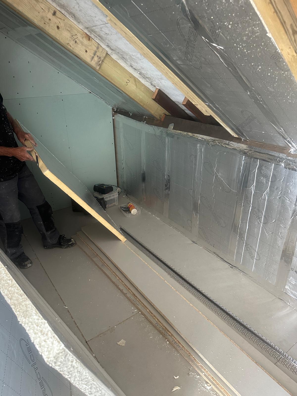 A person installs insulation panels and plasterboard in a partially finished loft space with building materials on the floor.