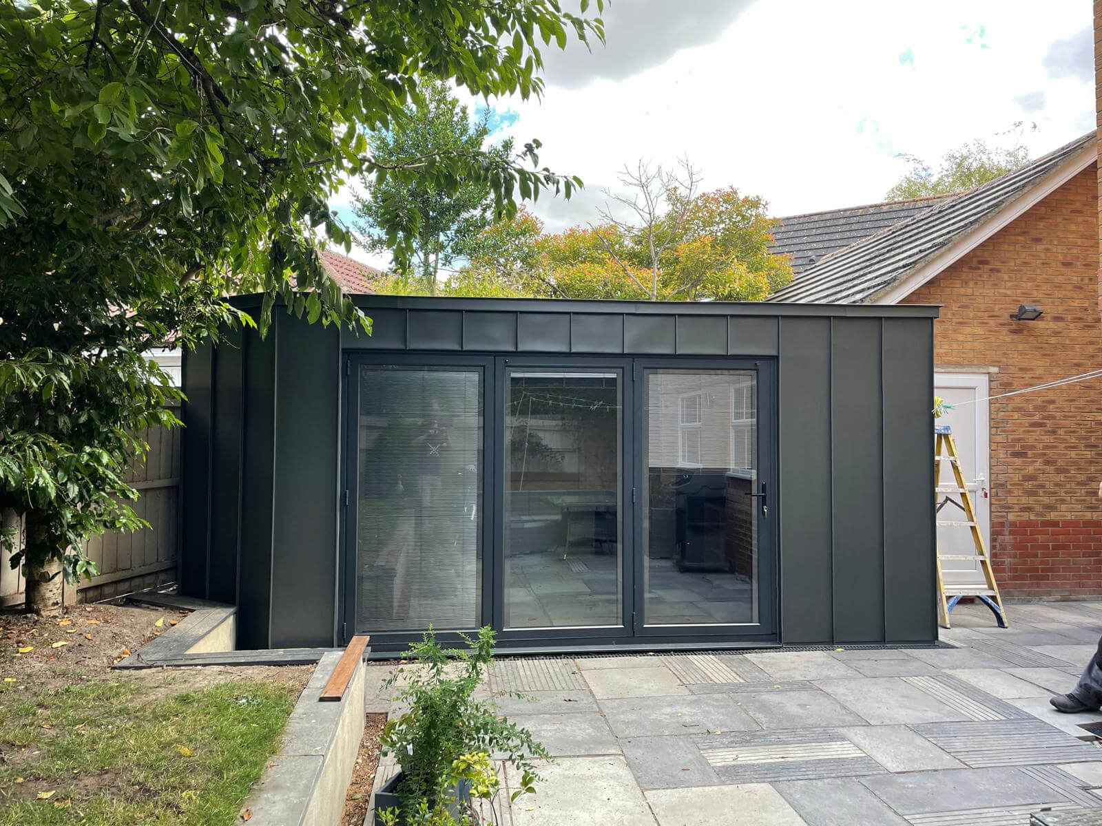 A modern black garden room with large glass sliding doors stands on a paved patio beside a brick house, where Sash Window Repair South London services are nearby and trees create a peaceful backdrop.