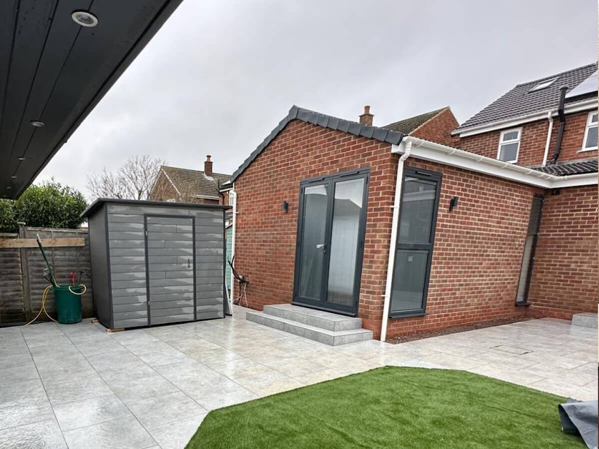 A modern brick outbuilding with glass doors and steps stands next to a grey storage shed, tiled patio, and artificial grass area under an overcast sky—ideal for those seeking Sash Window Repair South London expertise.