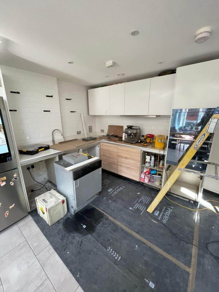 Modern kitchen under renovation with tools, ladder, and appliances. Cupboards are partially installed; floor is covered for protection.