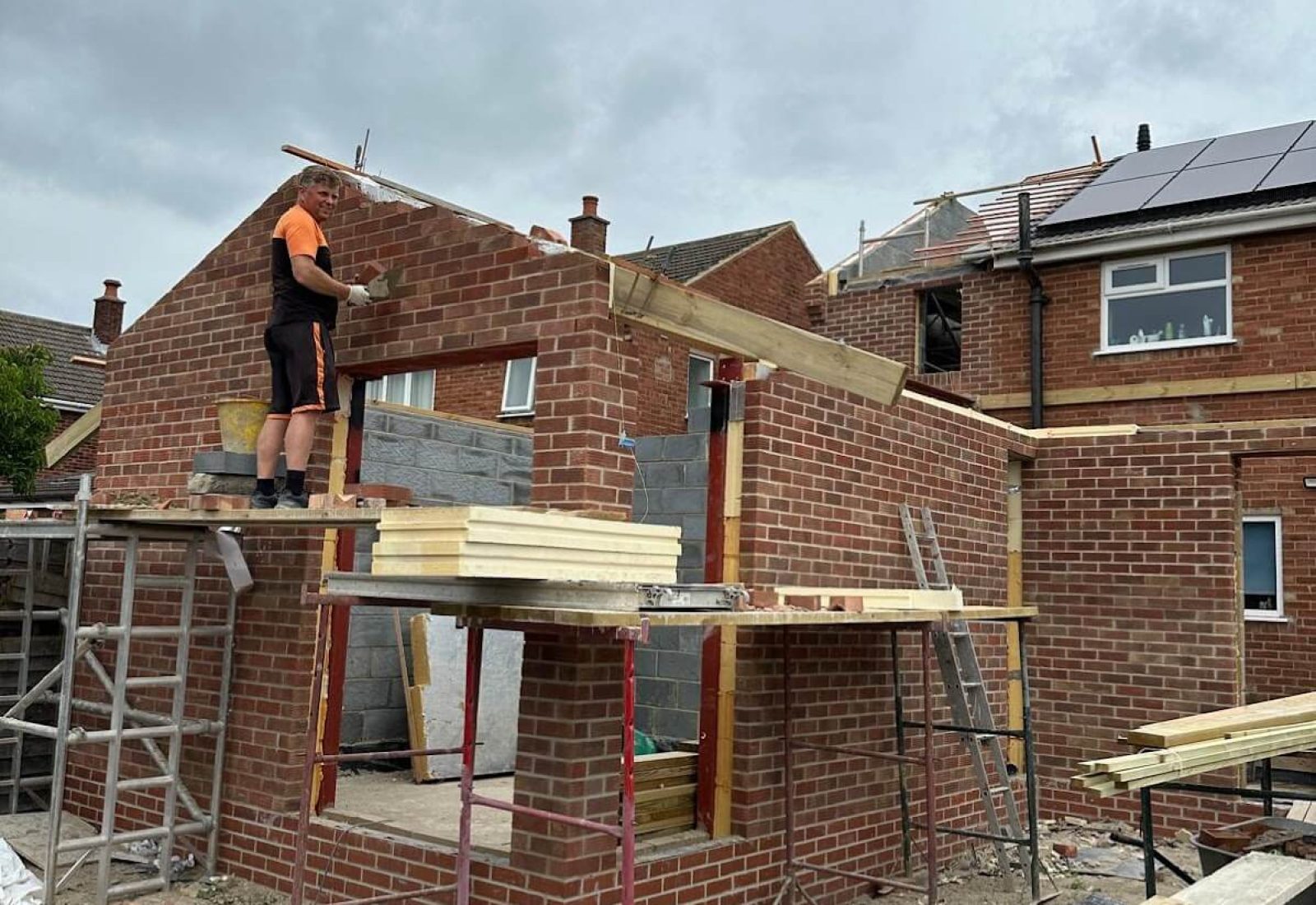 A construction worker stands on scaffolding while building a brick extension to a house, where sash window repair South London services may soon be needed. Wooden beams and building materials are scattered around the site.
