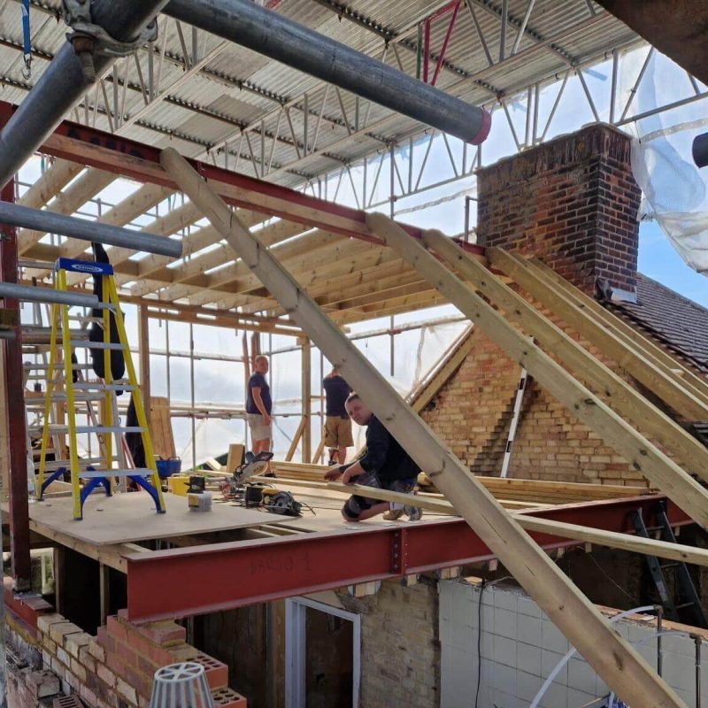 Several workers construct a timber roof frame on top of a partially demolished brick building, with scaffolding and tools visible around the site.