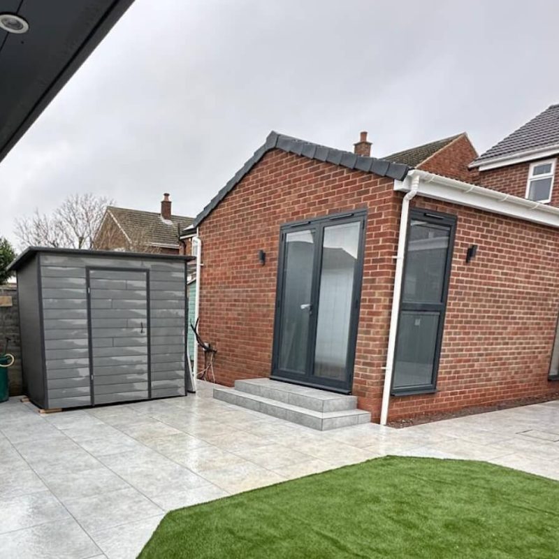 A modern brick outbuilding with glass doors and steps stands next to a grey storage shed, tiled patio, and artificial grass area under an overcast sky—ideal for those seeking Sash Window Repair South London expertise.