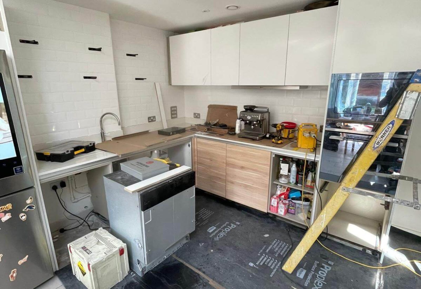 Modern kitchen under renovation with tools, ladder, and appliances. Cupboards are partially installed; floor is covered for protection.